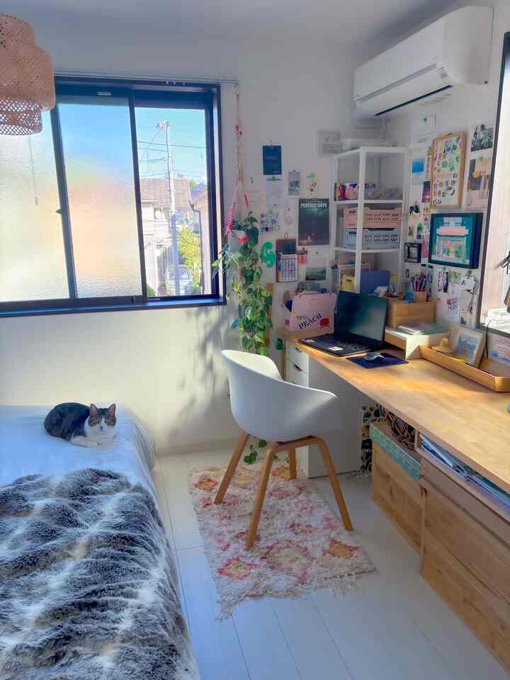 White-walled small workspace with beige wooden desk, a cat resting on bed, and natural light filling the cozy room