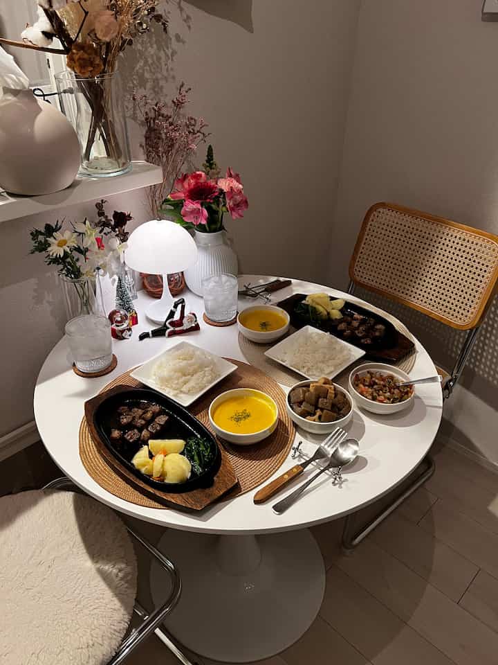 ナチュラルな木目調とホワイトの二人用ダイニングスペース、花瓶とテーブルランプが配置された雰囲気のあるインテリア