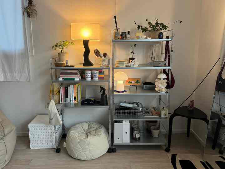 明るい白い壁と金属製の棚があるミニマルなリビング、黒いフロアランプとクリーム色のビーズクッションが配置された雰囲気のある空間