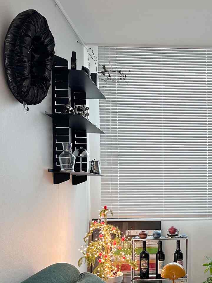 White-toned living room featuring blinds, black wall shelves, a wine rack, and a lit decorative plant creating a calm atmosphere
