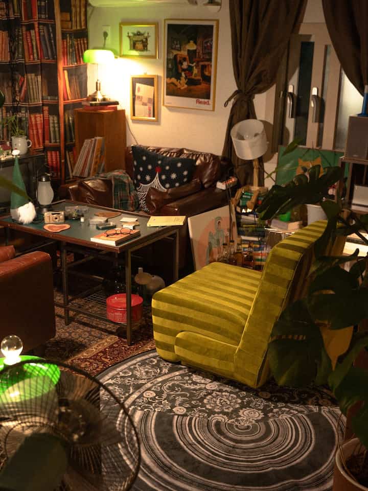 温かみのあるブラウンとオリーブグリーンのリビングルーム、ヴィンテージのレザーソファとグリーンのストライプチェア、パターンのラグと本棚が配置された雰囲気のあるインテリア