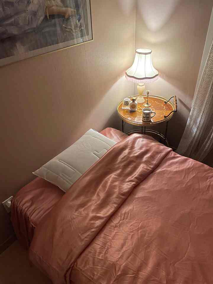 ピンクの寝具とベージュの壁が調和した居心地の良い寝室、横には上品なテーブルランプが置かれたサイドテーブルが配置されている