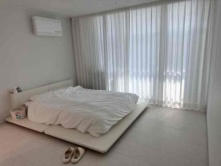 Bright white-toned bedroom featuring a low-profile bed and sheer white curtains in a clean, modern setting