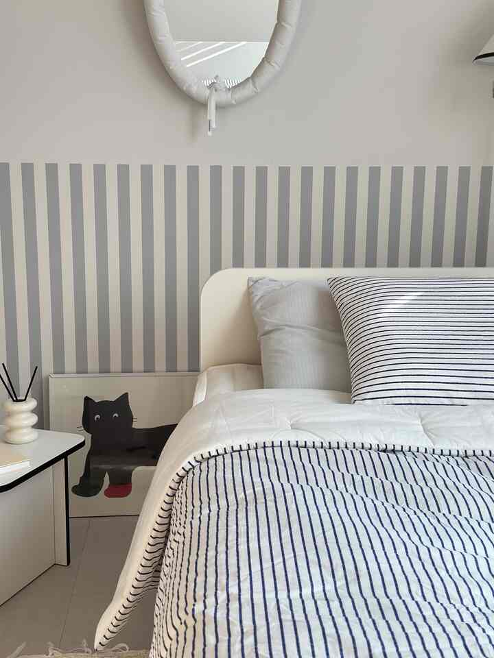 Modern bedroom in white and navy striped bedding featuring oval cushion mirror on wall and framed cat art on floor