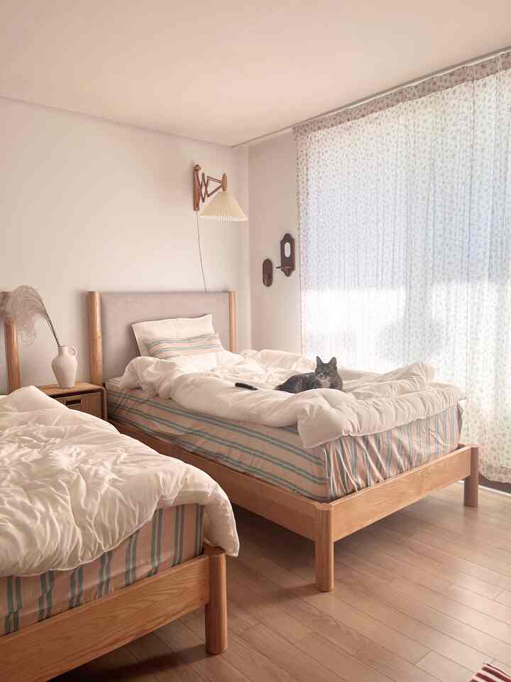 Natural wood-tone bed frames with white bedding and floral curtains in a two-person bedroom featuring a cat lying on one bed