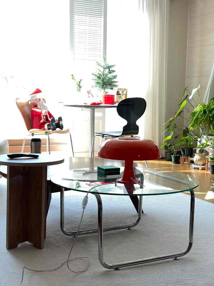 A modern living room with a white and black color scheme featuring a glass coffee table, red table lamp, and various plants in a bright natural setting