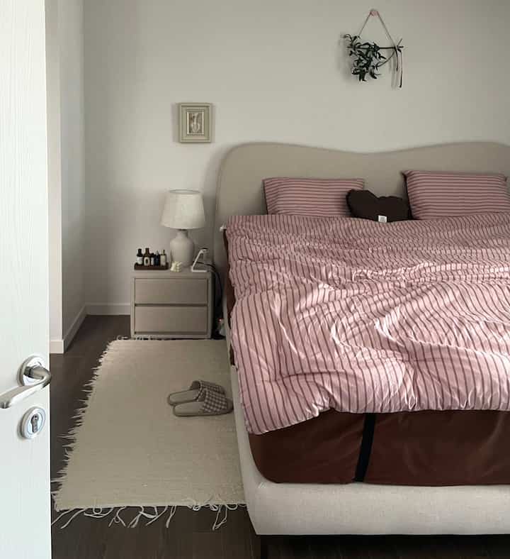 ベージュとホワイトの色調の寝室、ベッドとナイトテーブル、ピンクのストライプの寝具が配置された落ち着いた雰囲気のインテリア