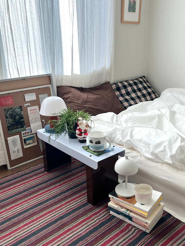 白と茶色のトーンの寝室、チェック柄の枕と掛け布団カバー、小さなテーブルランプと植物が配置された落ち着いた雰囲気のインテリア