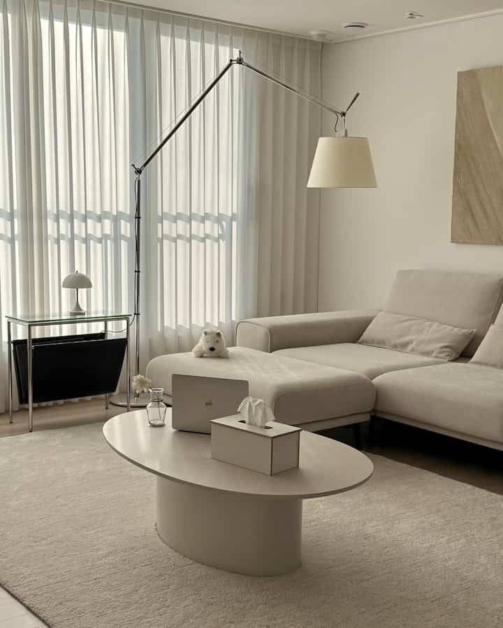 ベージュと白を基調としたリビングルーム、広々としたセクショナルソファとオーバルのコーヒーテーブル、カーテンとフロアランプが配置されたモダンで自然な雰囲気のインテリア