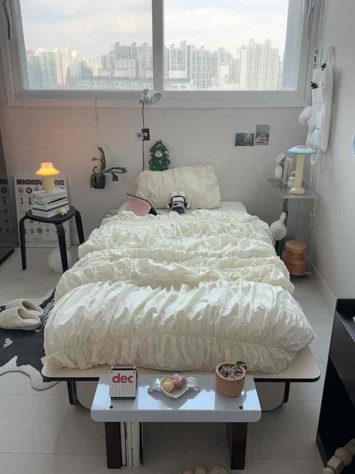 アイボリーと白を基調とした寝室で、中央に置かれたベッドと隣の小さなデスク、植物が調和する印象的な空間