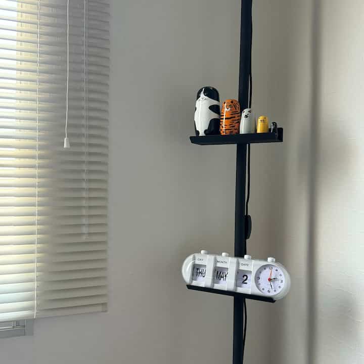 白を基調としたワンルーム空間で、左側にアルミブラインド、中央のテンションロッド棚には時計とマトリョーシカ人形が配置されたシンプルなインテリア