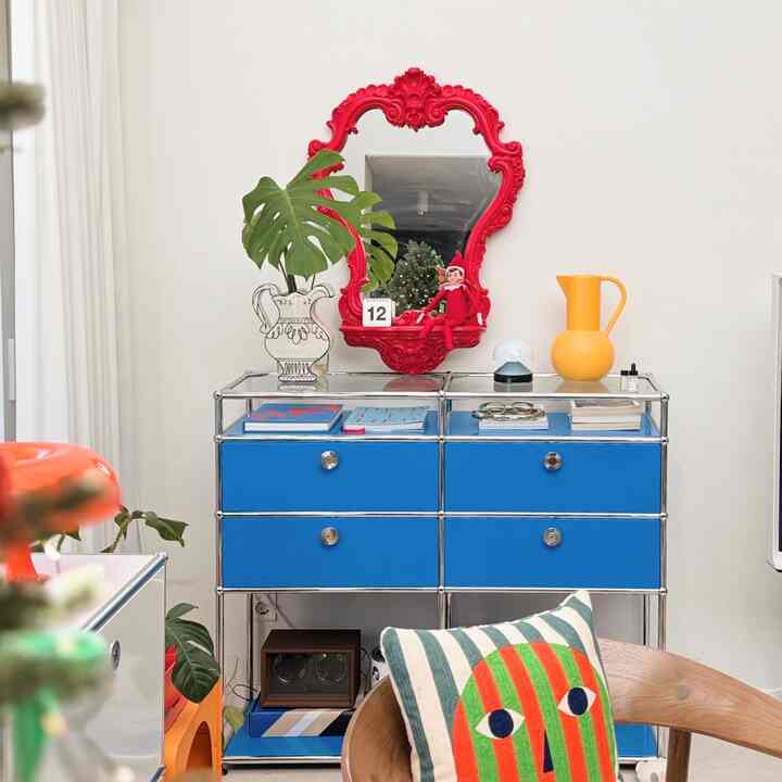 白い壁と青いモジュール収納のリビング、鮮やかな赤い装飾鏡と多様な小物が配された雰囲気のあるインテリア