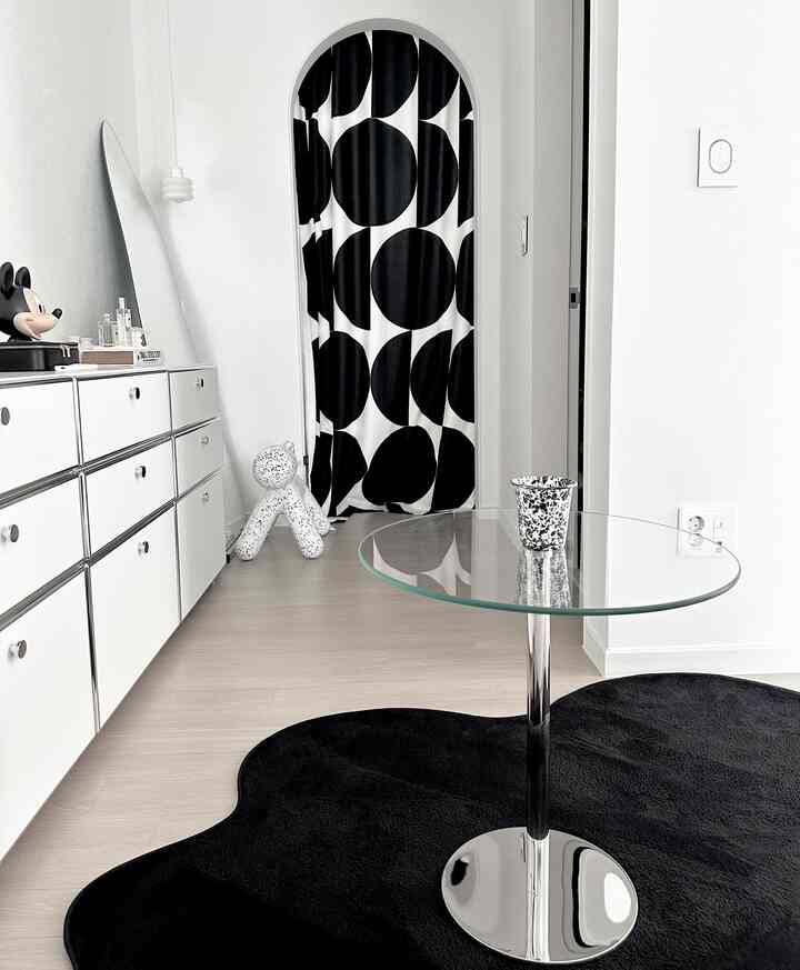 モノトーンでミニマルな小さなリビング、黒いラグと丸いガラステーブル、モノクロの柄のカーテンが調和した空間