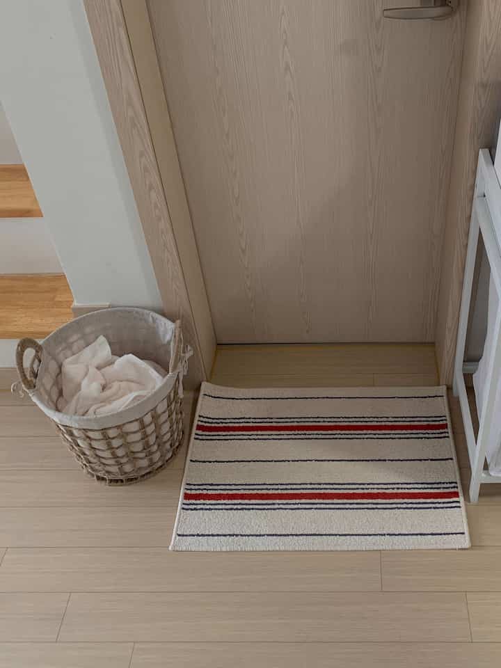 ベージュと白のトーンの玄関、木製ドアと床、ランドリーバスケットとストライプのバスマットが配置された温かみのある空間