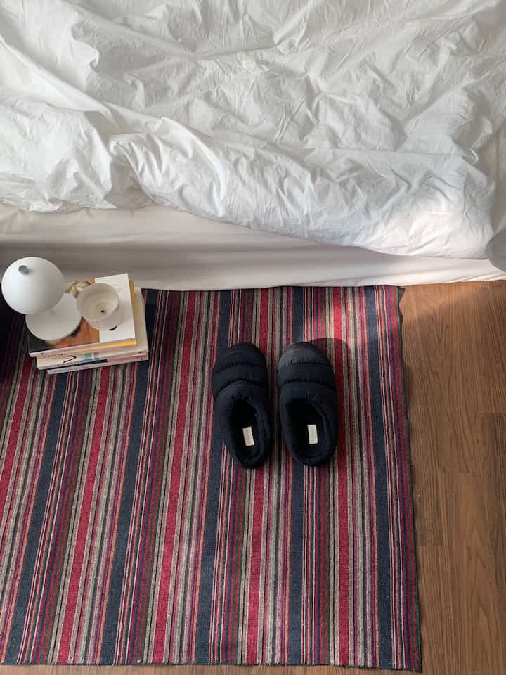 白い寝具と茶色い床、ストライプのラグの上にパッド入りルームシューズがある寝室の雰囲気あるインテリア