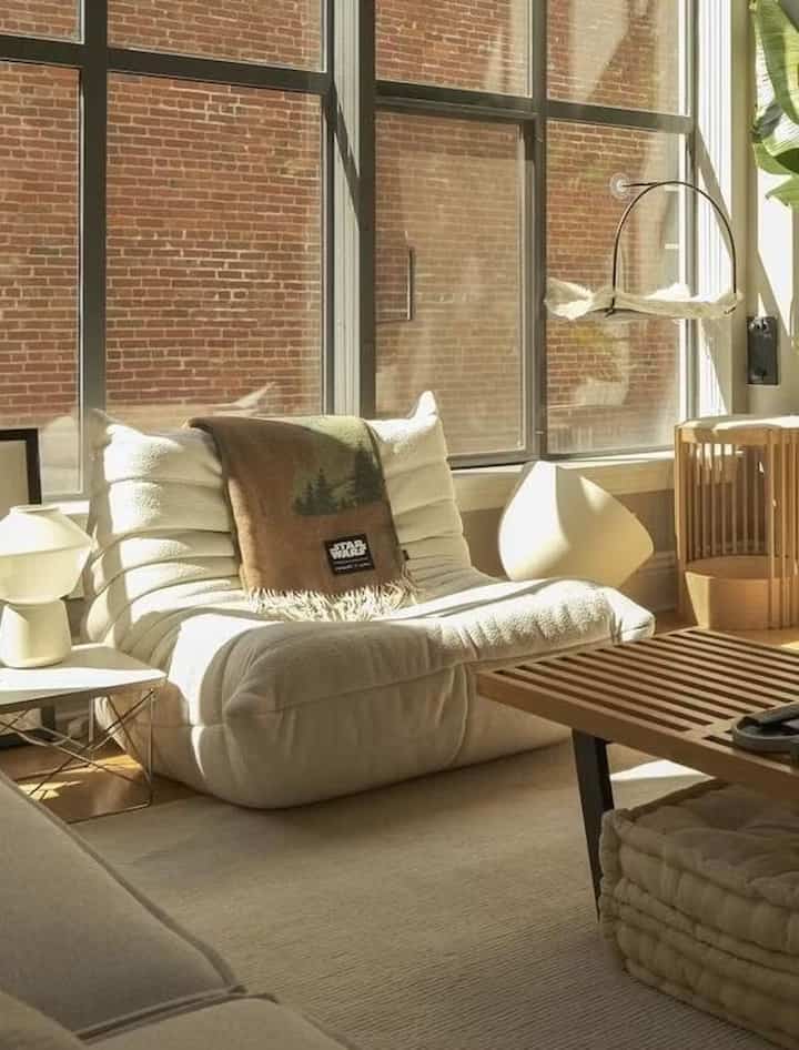 Bright beige and white toned living room featuring a cozy armchair near large windows and a wooden coffee table in a warm atmosphere