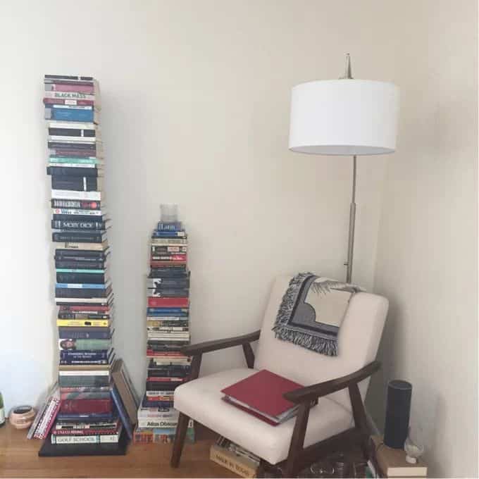 Beige-toned living room corner featuring an armchair, floor lamp, and stacks of books arranged in corner creating a cozy reading nook