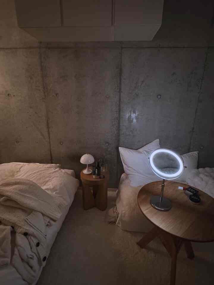 Small bedroom with gray concrete wall and white bedding, featuring natural wood tables and simple table lamps in a cozy setting
