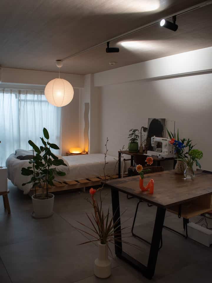 Natural tone studio apartment with wood ceiling, featuring a bed, dining table, and various plants in a calm atmosphere