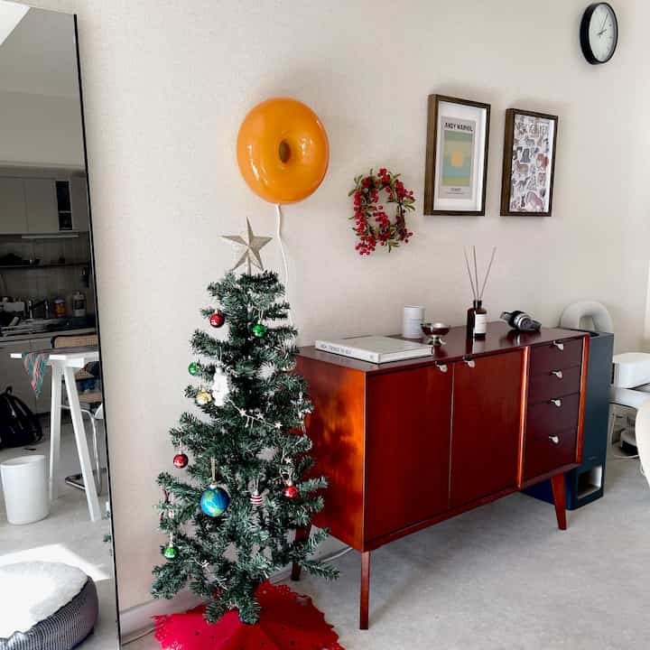 白い壁と茶色の木製家具が配置された寝室、クリスマスツリーとオレンジ色の壁掛け照明が印象的なナチュラルモダンなインテリア