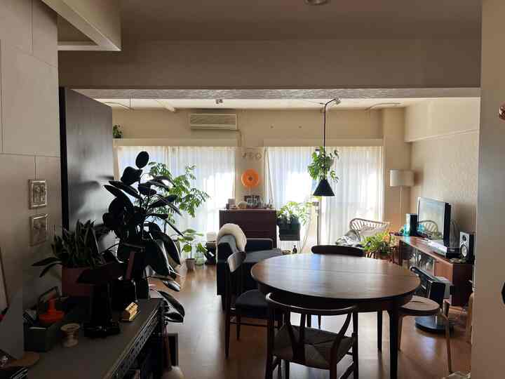 ベージュの壁と木目調家具が配置されたリビング兼ダイニングルーム、中央に丸いダイニングテーブルとソファ、植物がある自然な雰囲気の空間