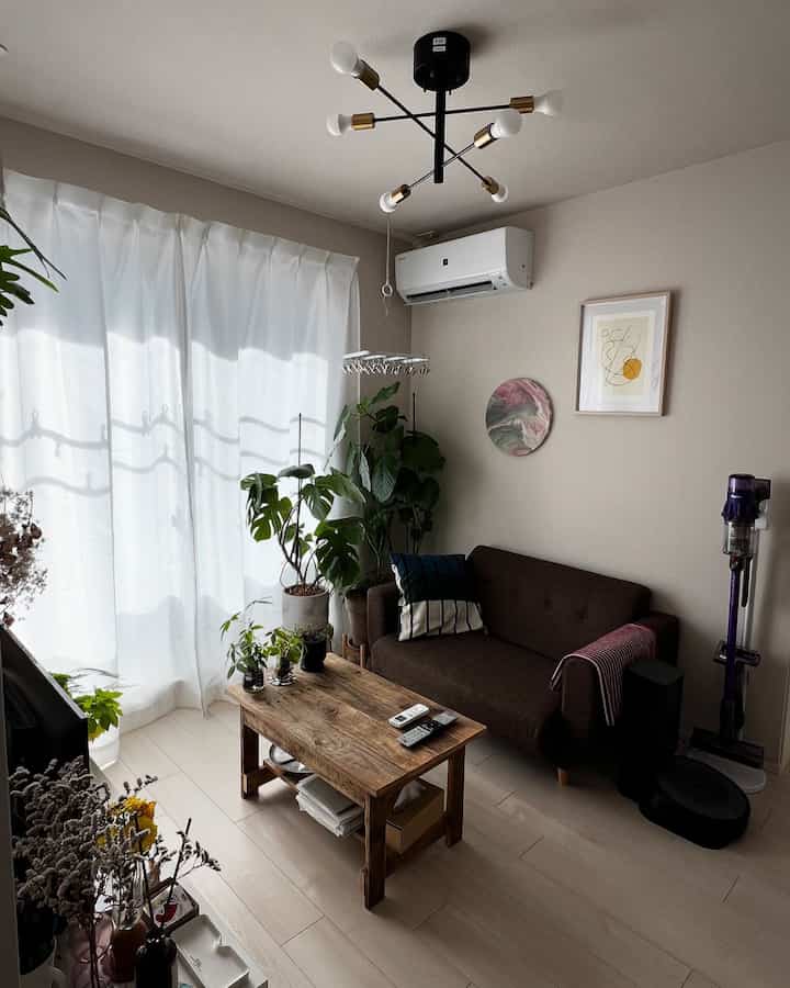 ナチュラルな木目調家具と白いカーテンが調和したコンパクトなリビング、ダークブラウンのソファと観葉植物が配置されたシンプルなインテリア