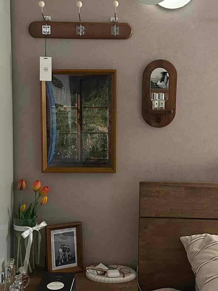 Natural wood tone and beige bedroom wall featuring a wooden mirror, picture frames, nightstand with vase and decor, creating a cozy space