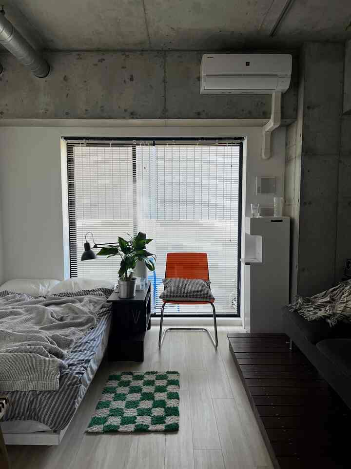 白とダークブラウンのトーンのワンルーム寝室、オレンジのオフィスチェアと植物、ベッドとダークソファが整然と配置されたモダンな空間