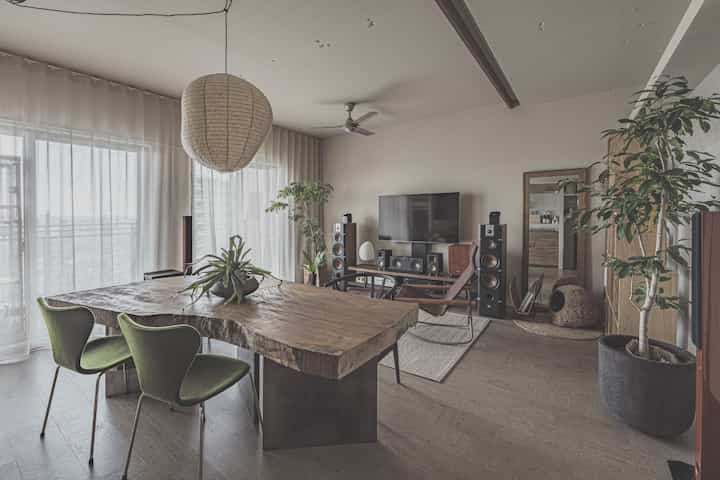 ナチュラルトーンのリビング、広い一枚板のダイニングテーブルとグリーンチェア、多数のオーディオスピーカーが配置された雰囲気のあるインテリア