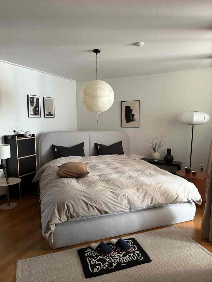 ベージュと黒のトーンの寝室、丸い和紙の照明とモダンなベッド、ラグ、額縁が配置された落ち着いた自然なインテリア