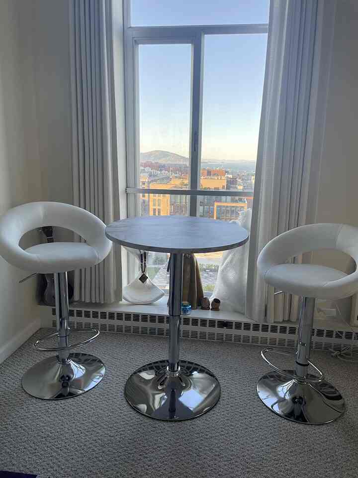 Modern minimalist home bar featuring two white bar stools and a round adjustable pub table positioned in front of a scenic window view