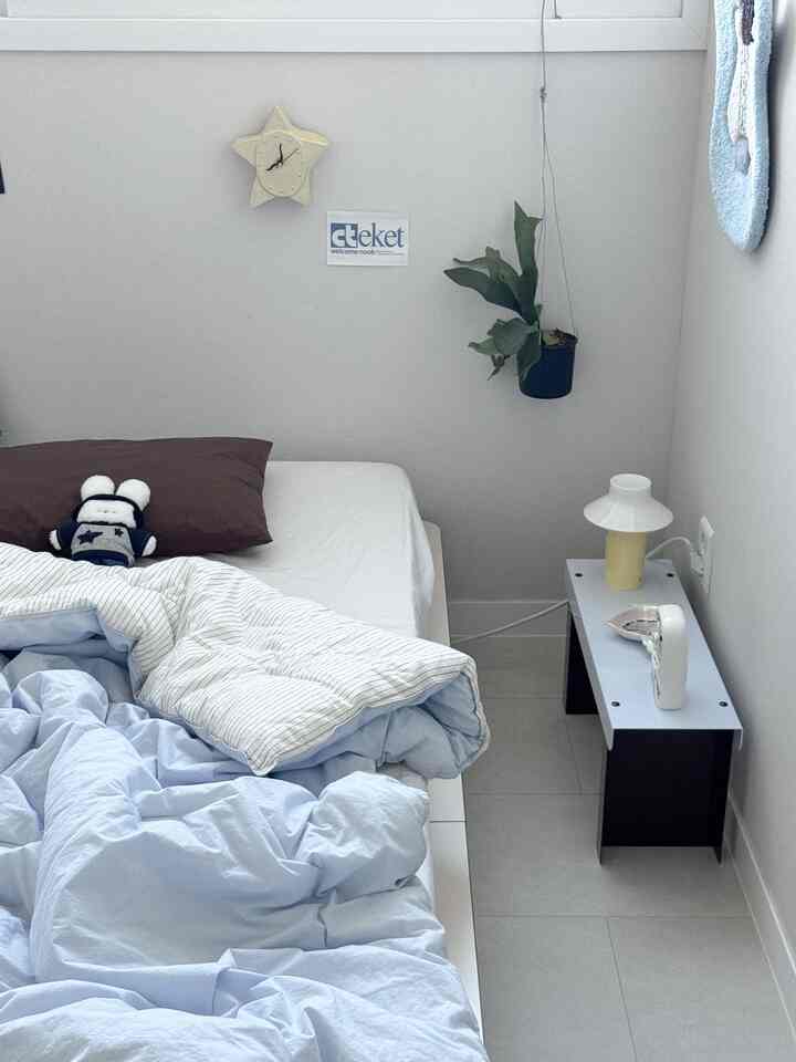 白とライトブルーの色調の寝室、シンプルなベッドとナイトテーブル、観葉植物が配置された落ち着いた空間