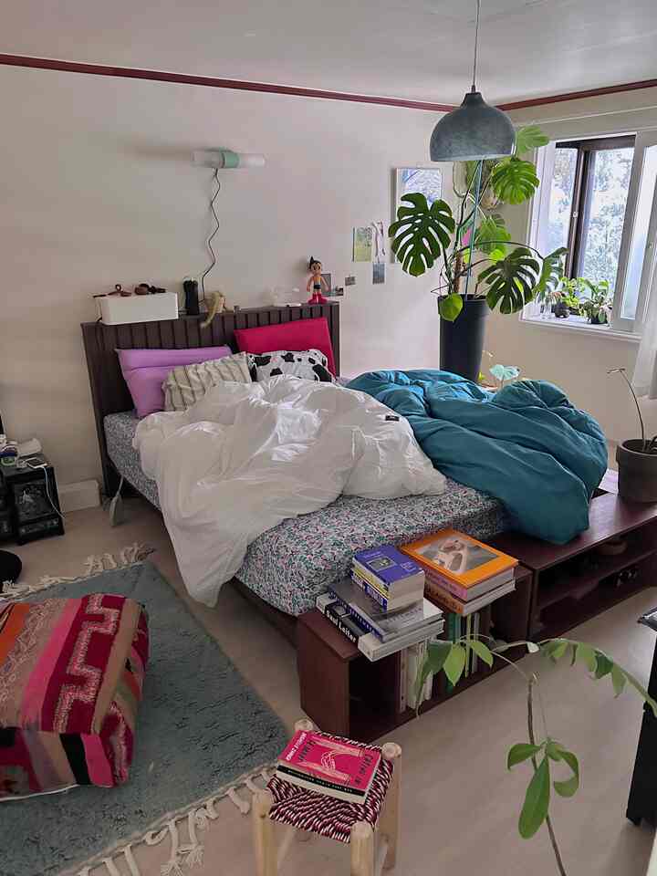 ナチュラルトーンの寝室、中央にベッドと白・ターコイズの掛け布団カバー、大きなモンステラの植物が窓際にある落ち着いた空間