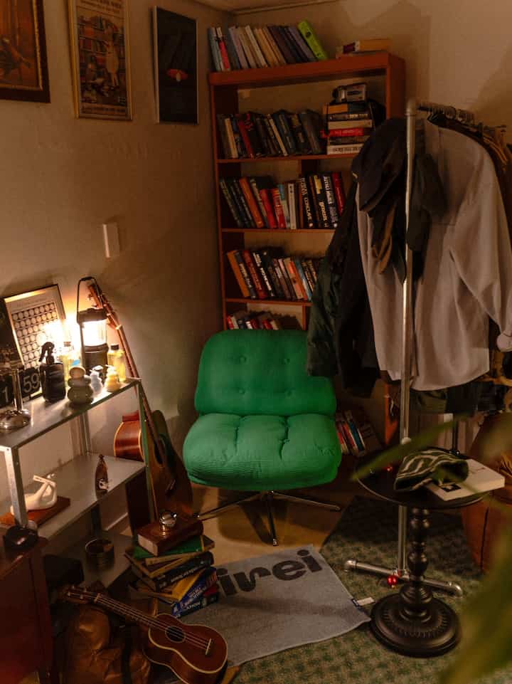 暖かいベージュ色調の小さなリビングスペース、グリーンのアームチェア、本棚、コートラック、テーブルランプが配置された雰囲気のある読書空間