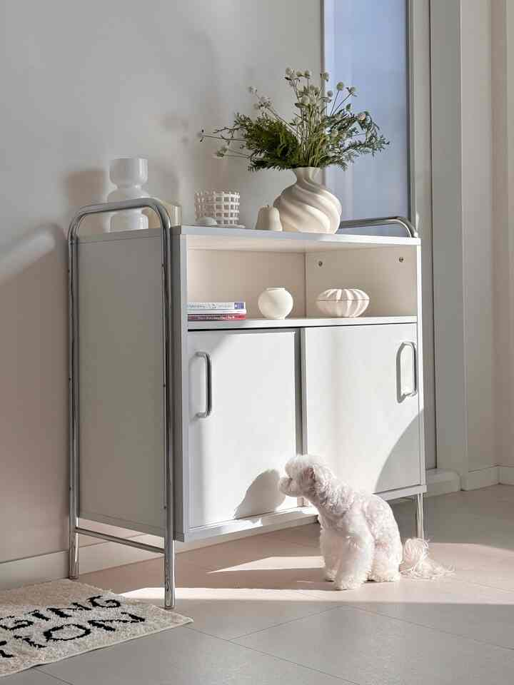 明るいホワイトとシルバーのトーンのキャビネットが配置された空間、上部には花瓶とオブジェがあり、下部には小さな白い犬が座っている温かみのあるインテリア