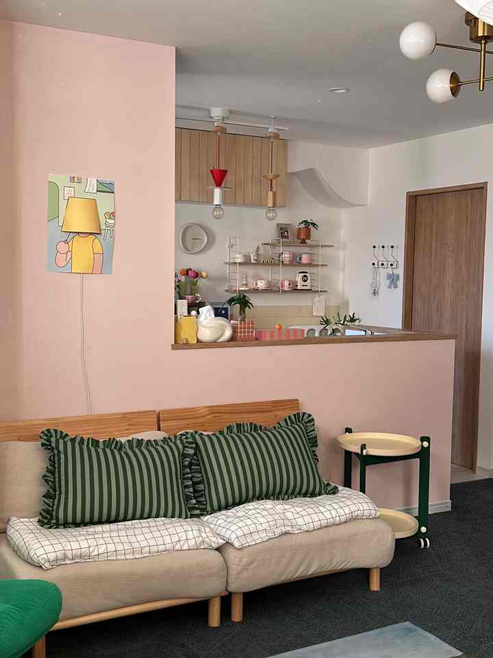 パステルピンクの壁とベージュのソファがある韓国インテリアのリビング、緑のストライプクッションと木目調の棚が配置された雰囲気のある空間