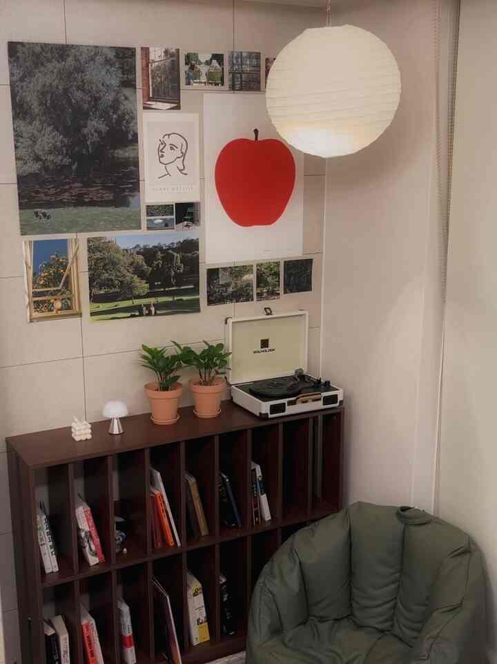 ナチュラルな色調の小さな書斎スペース、グリーンのビーズクッションとダークブラウンの本棚、多彩な額装アートと柔らかなライトが配置された落ち着いたインテリア