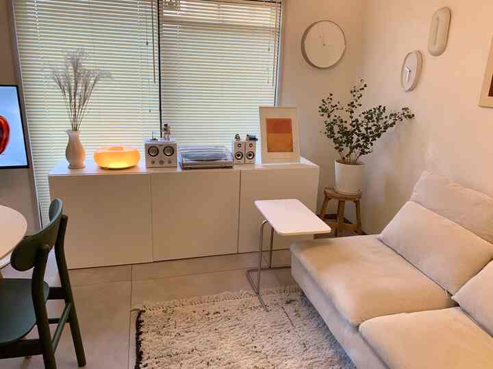 A white and natural toned living room featuring a beige sofa, white cabinet, plant, and poster creating a cozy atmosphere