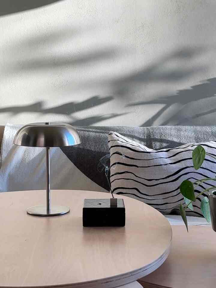 ベージュの丸い木製リビングテーブルにシルバーのテーブルランプと黒の香立が配置され、ストライプのクッションと柔らかな壁の影が雰囲気のあるインテリアを演出