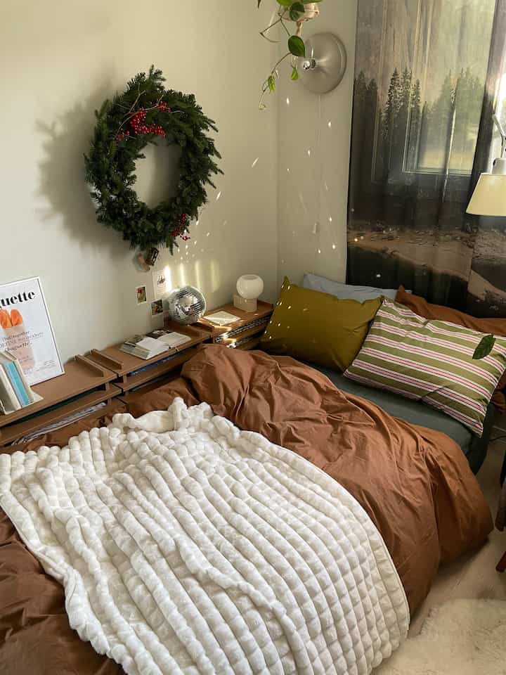 ブラウンとホワイトのトーンの寝室、壁に飾られたリースと複数のクッション、温かみのあるブランケットがある雰囲気のあるインテリア