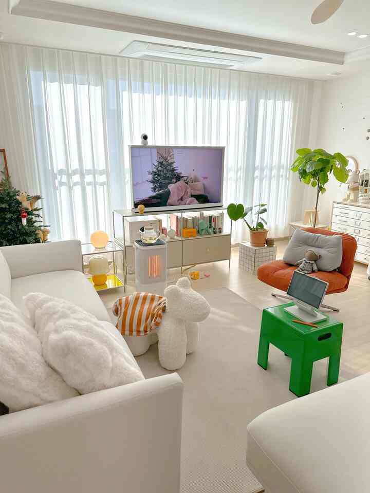 A bright living room dominated by white and accented with orange, featuring large sofas, a TV stand, plants, and mid-century modern style elements