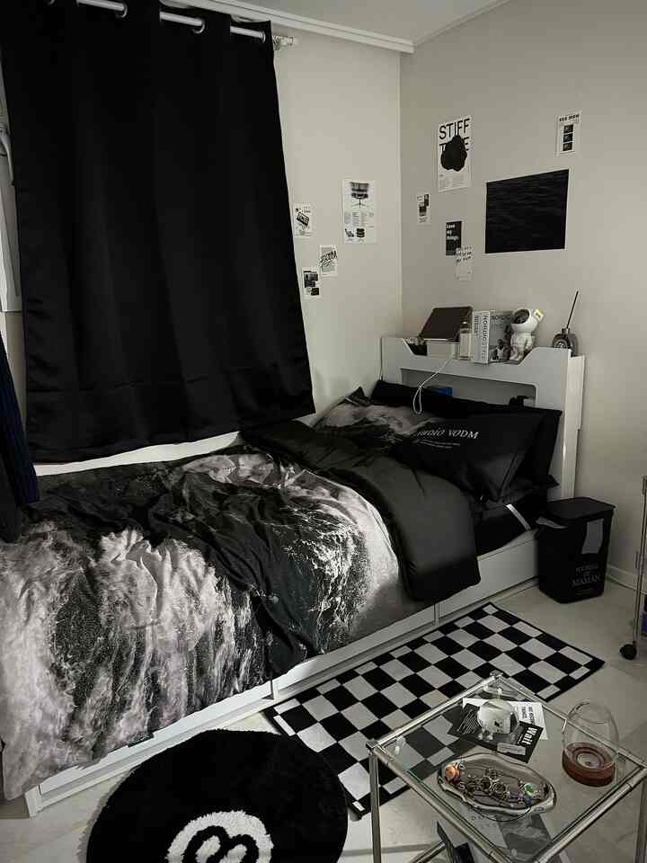 Monotone black and gray bedroom featuring a bed with ocean-themed duvet cover, black blackout curtain, and multiple posters on the wall in a modern setting