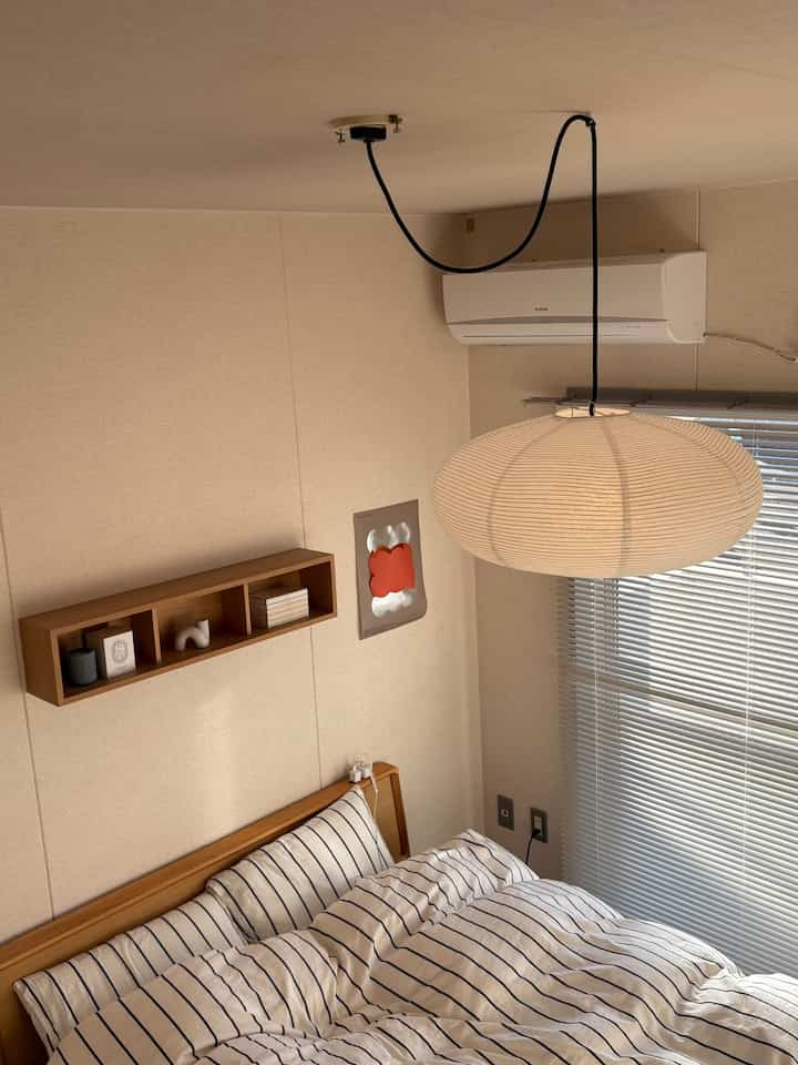 ベージュと白のナチュラルカラーの寝室、木製の壁掛け棚とストライプの布団カバーが配置された落ち着いた空間