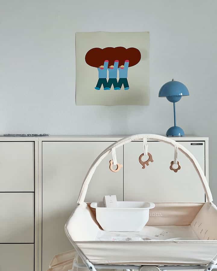 ホワイトとナチュラルトーンの赤ちゃん部屋、オムツ交換台とブルームードランプ、アートポスターが配置された清潔感のあるインテリア