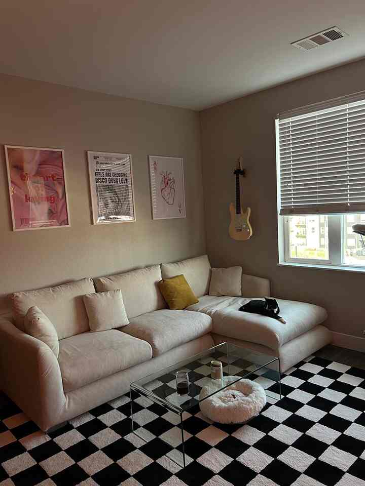 A beige and white toned living room featuring a cream sectional sofa, transparent glass coffee table, black and white checkered rug, and a cat lounging on the sofa, creating a cozy atmosphere