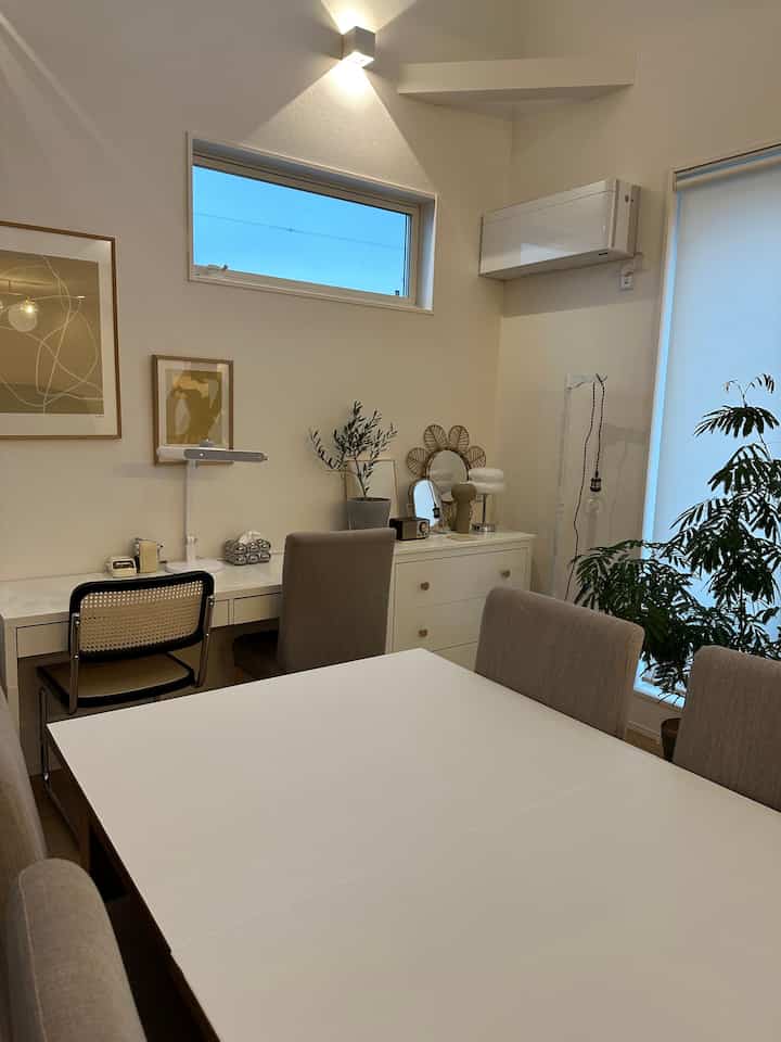 A white and beige toned dining room featuring a white dining table, desk, dresser, and natural plants, creating a calm and cozy atmosphere
