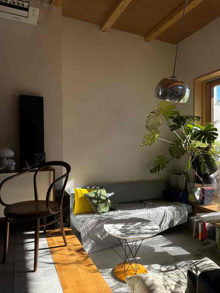 ナチュラルカラーと白い壁のリビング、ソファに黄色のクッション、丸いコーヒーテーブルと大きな植物が配置された印象的な空間