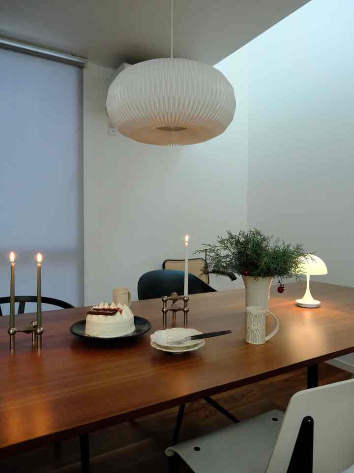 白い壁と茶色の木製ダイニングテーブルがあるシンプルでナチュラルなダイニング、テーブルにはキャンドルやケーキ、照明が配置されている