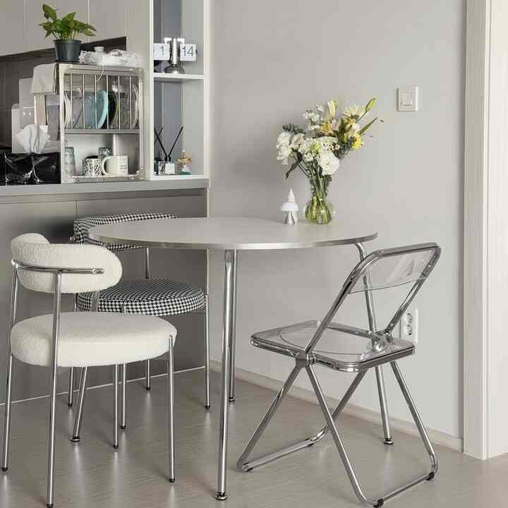 白とシルバーのトーンのダイニングルーム、透明な折りたたみ椅子と花瓶が配置されたモダンな食卓空間