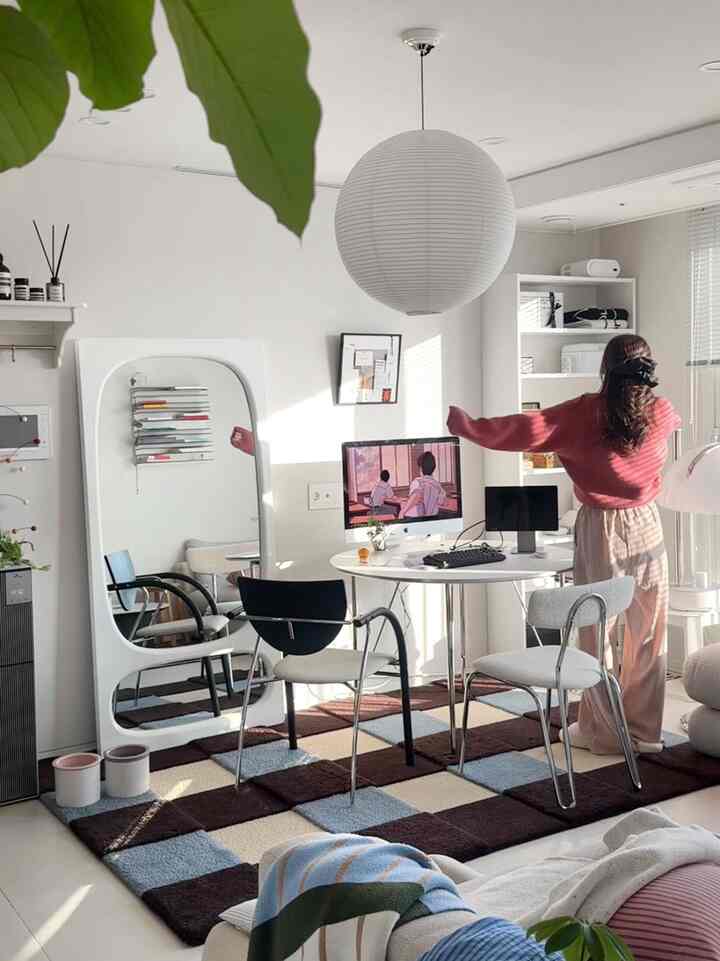 白と茶色のトーンでまとめられたリビング、丸いデスクにコンピューターとチェック柄のラグが配置されたミニマルで明るい空間